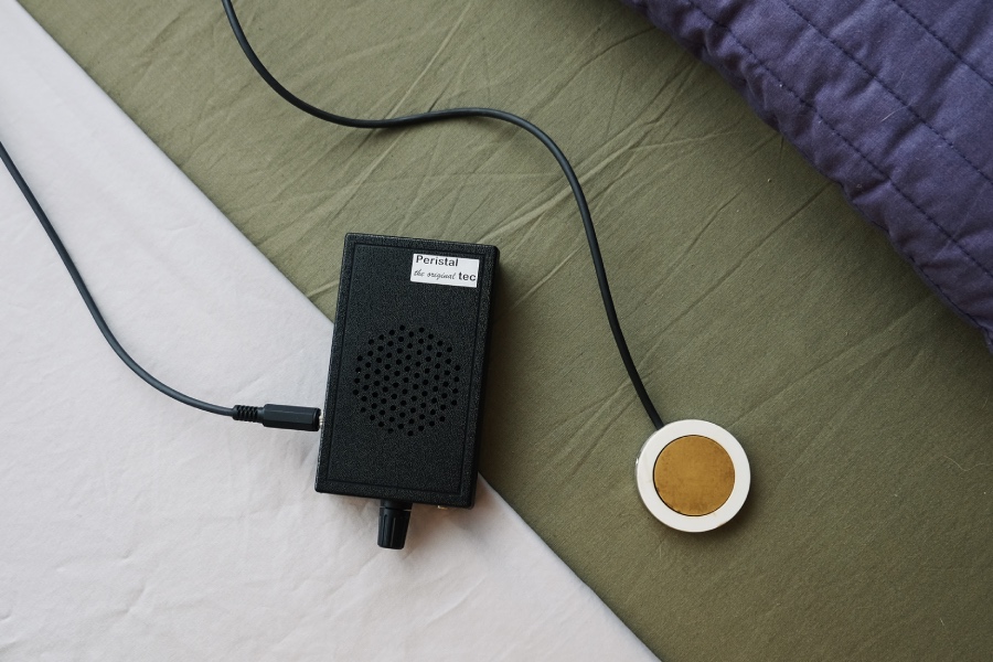 Small black rectangular electronic device with a perforated grille, connected by a cord to a round white and brown sensor pad on a bedspread.